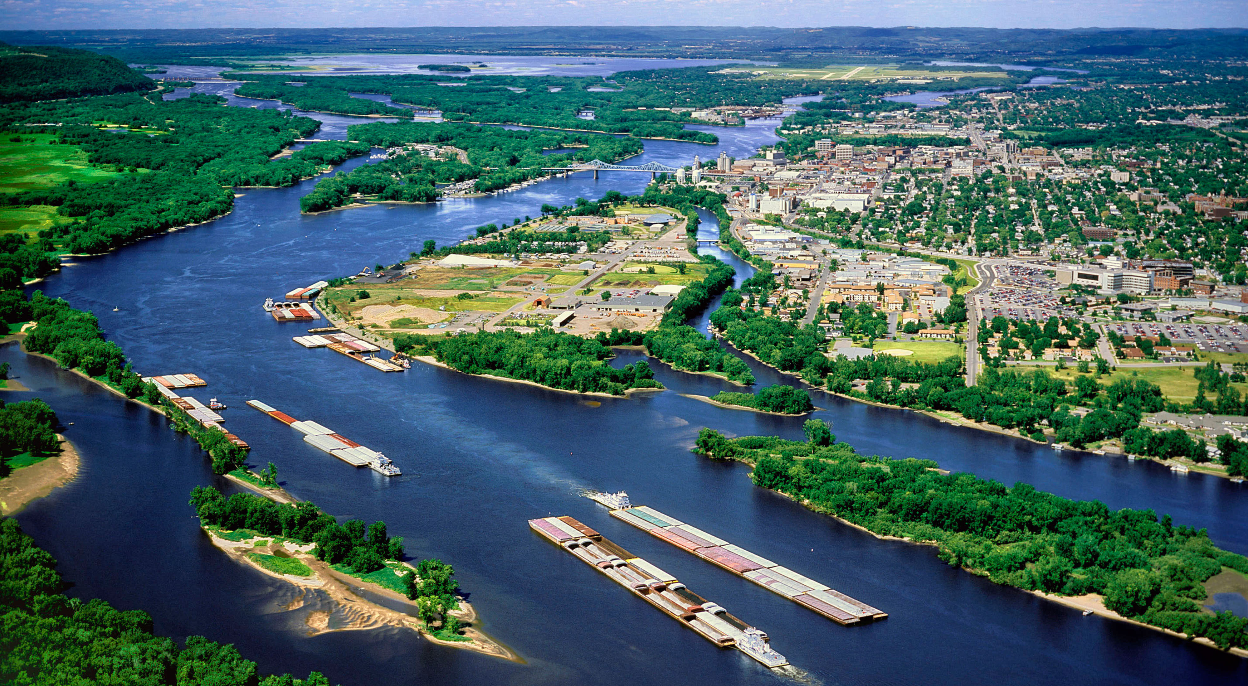 Vista aérea de barcaças no rio Mississippi e suas curvas contornando áreas povoadas e florestas.